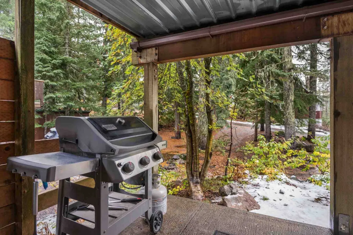 An exterior view of the deck featuring a grill with the surrounding forest in full view. The setup suggests outdoor dining and barbecuing opportunities, with a pathway leading into the trees and room for communal gatherings. The relaxed mood hints at summer grilling parties or morning coffees outdoors.