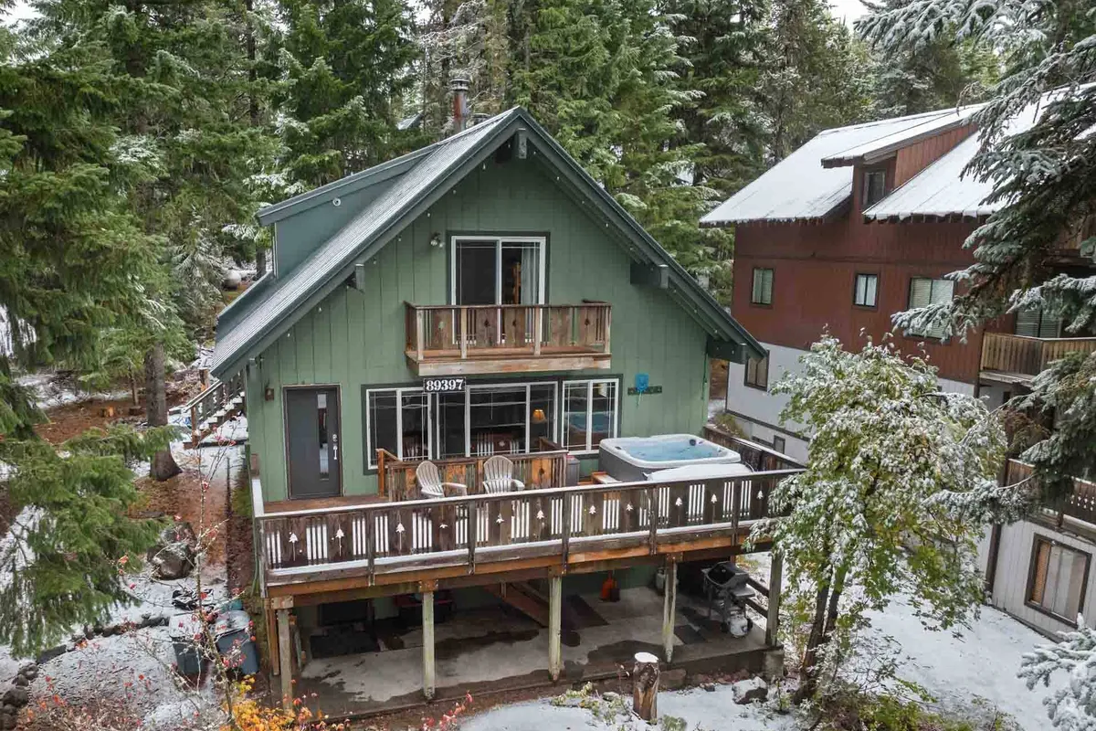 The image features the green exterior of the Mt. Hood Chalet, nestled among snow-covered trees. The spacious balcony is furnished with chairs, inviting guests to relax and soak in the tranquil mountain setting, while a private hot tub offers a cozy spot to watch the snowfall or stargaze at night.