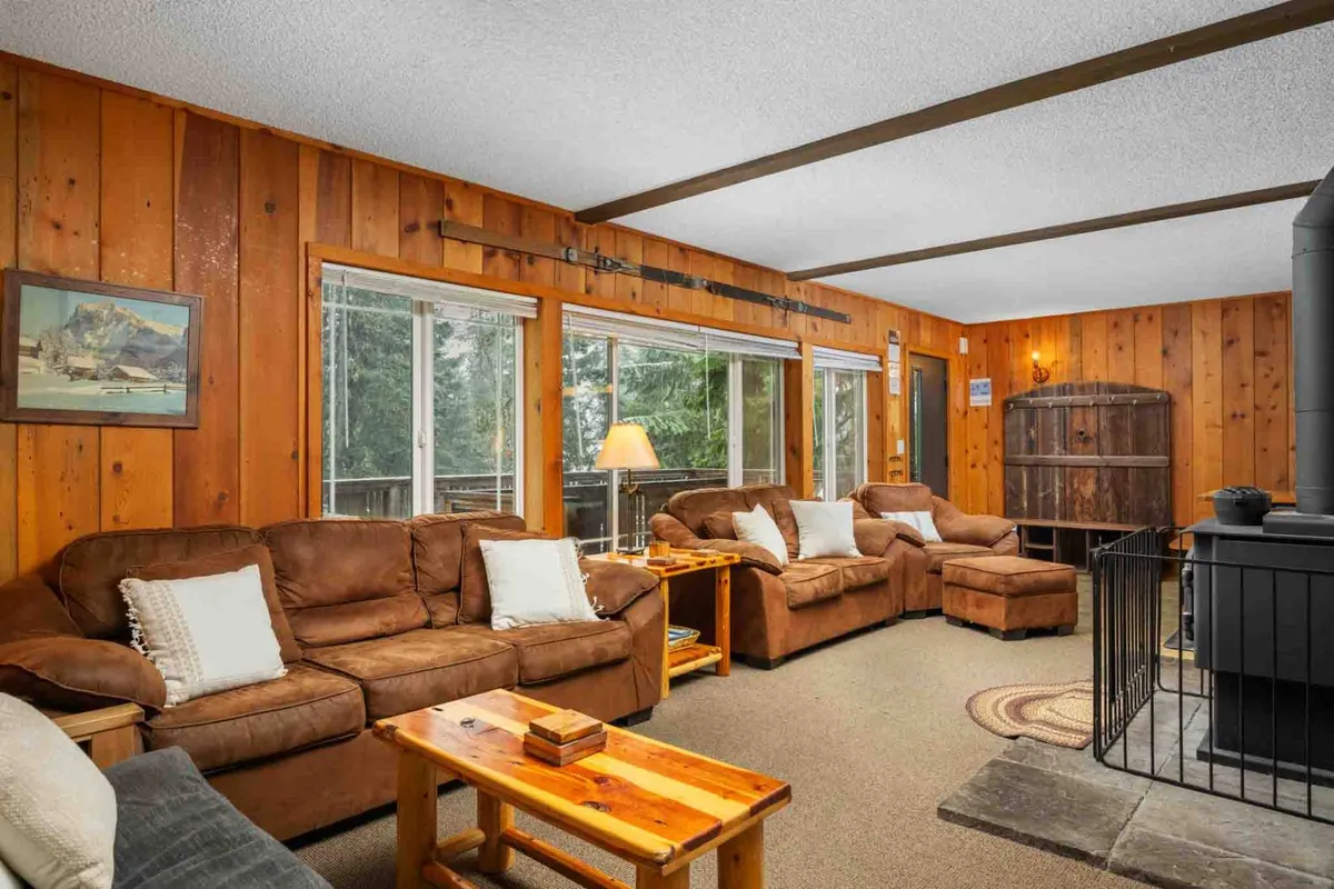 A warm, welcoming living area featuring comfortable sofas arranged around a wooden coffee table, wooden walls that enhance the lodge-inspired ambiance, a cozy fireplace, and large windows that fill the space with natural light and showcase views of the surrounding forest. The scene evokes togetherness, relaxation, and joyful gatherings.