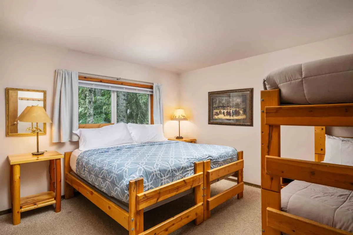 A cozy bedroom featuring a queen bed with patterned bedding, a bunk bed, a lamp on the bedside table, and framed artwork on the wall. Sunlight filters in through a nearby window, creating a welcoming, family-friendly atmosphere ideal for both adults and children.