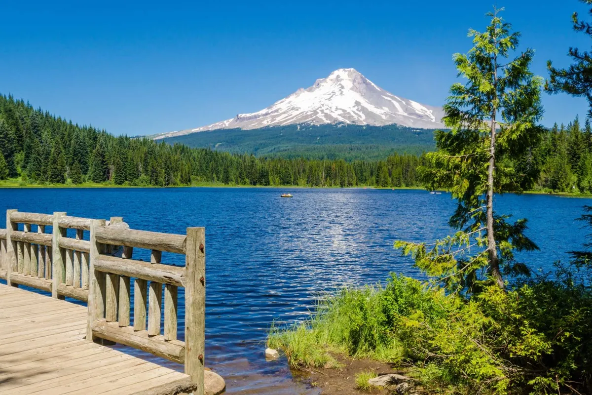 A serene view of a calm lake with a wooden pier, lush greenery, and a distant mountain peak. The landscape is tranquil and inviting, ideal for activities like hiking, paddling, fishing, or picnicking in the warmer months.