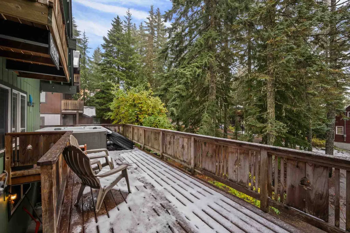 A cozy wooden deck nestled among towering trees, featuring a private hot tub and Adirondack chairs. The surrounding forest creates a sense of seclusion and tranquility, while the deck offers ample space for gatherings or relaxation. Snow blankets the ground, evoking a winter retreat atmosphere, but the setting is equally inviting for summer mornings or evening drinks outside.