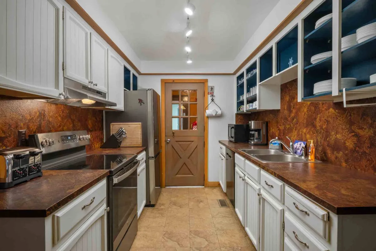 The image showcases a warm, inviting kitchen with gleaming stainless steel appliances, wooden cabinets, open shelving filled with dishes, and a spacious kitchen island. The setup invites group cooking and socializing, capturing the heart of a well-equipped main kitchen ideal for family gatherings or meal preparation for larger groups.