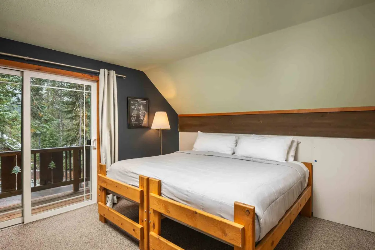 A tranquil bedroom featuring a comfortable bed positioned near a sliding glass door, a bedside lamp, and wall art. The view outside the door offers a calming natural backdrop, suggesting a peaceful environment for rest and relaxation.