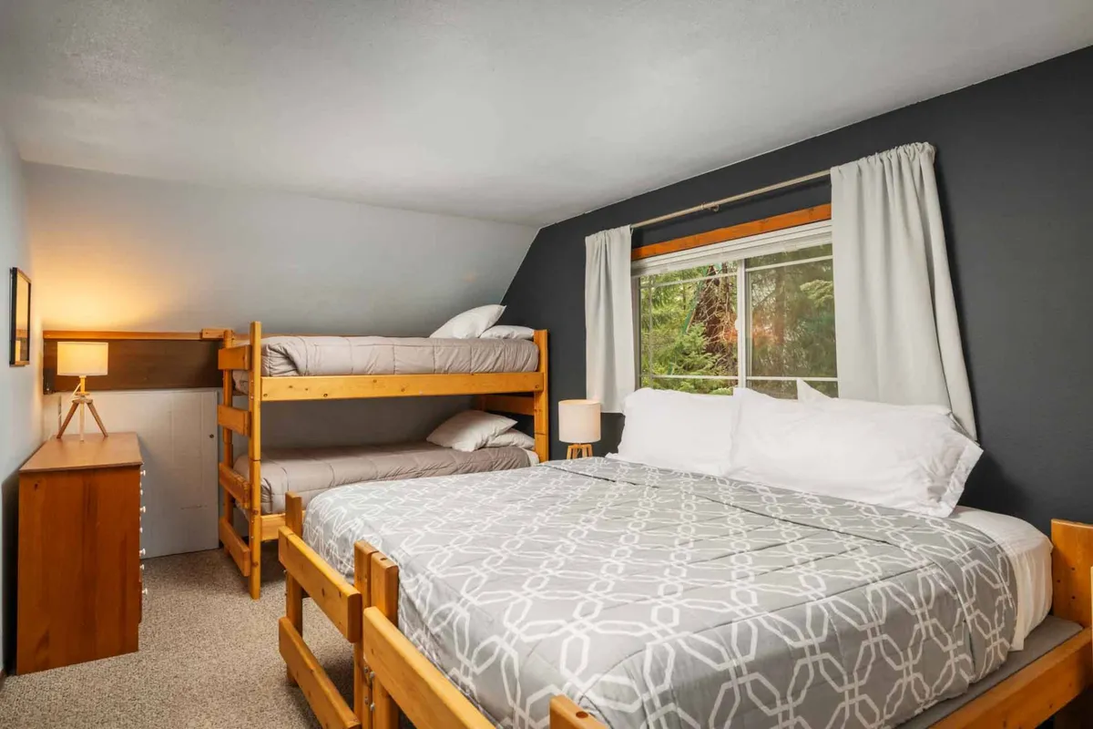 A spacious, light-filled room with a set of bunk beds and a queen bed, complemented by a window allowing natural light, a lamp on a bedside table, and a wooden dresser. The setup invites both relaxation and playful moments for families or groups.