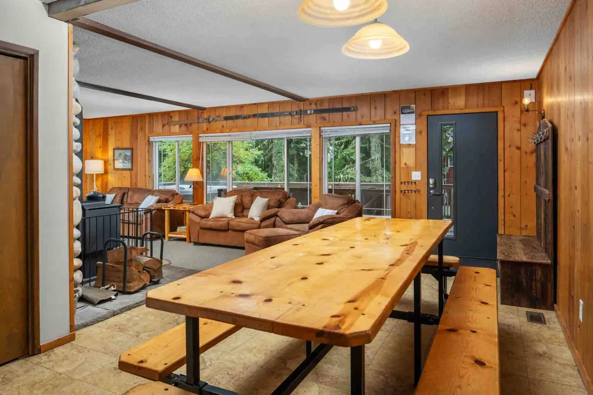 An open concept space where a dining table sits adjacent to the cozy living area, both illuminated by natural light streaming through large windows. Warm wooden paneling and inviting décor create the ideal setting for shared meals, lively conversation, and friendly gatherings.